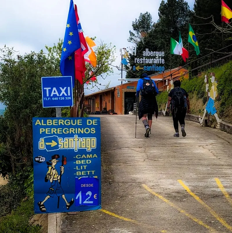 Two hikers walk up a gravel path towards a restaurant with flags and a sign for a pilgrims' hostel.