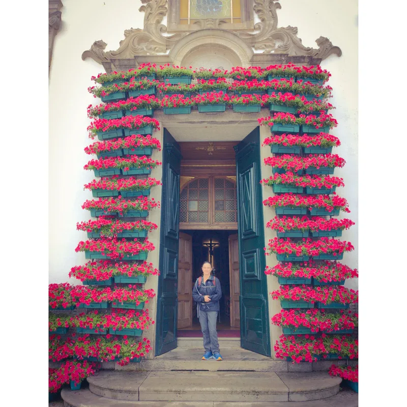 A person stands at a door surrounded by flowers in green planters, with open wooden doors revealing an interior.
