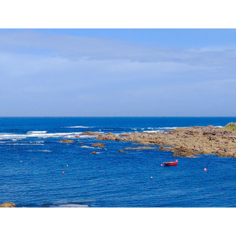 Calm blue ocean with rocky shoreline and a red boat near the shore, under a clear blue sky.