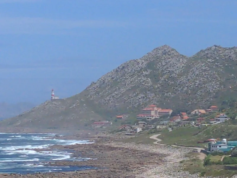 Coastal landscape with rocky shore, waves, a lighthouse on a hill, and buildings nestled among green hills.