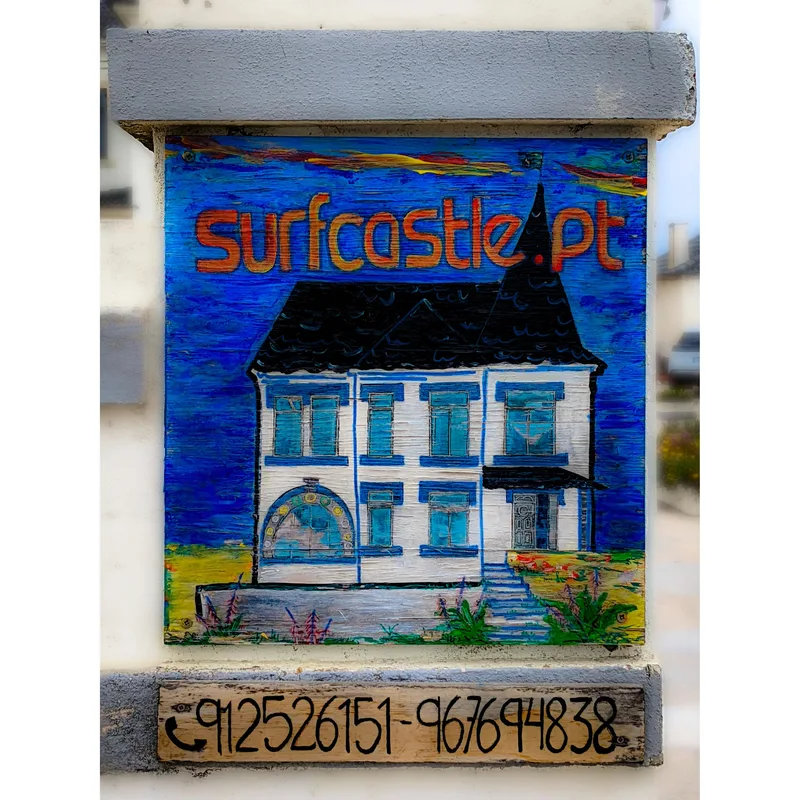 A colorful sign featuring an illustration of a white house with a black roof, labeled "surfcastle.pt" and contact numbers.