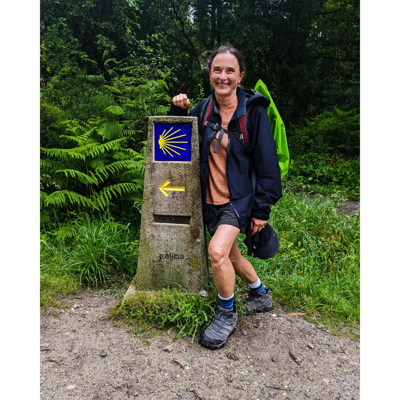 A woman stands next to a stone trail marker with a yellow arrow and a blue scallop shell design in a forested area.