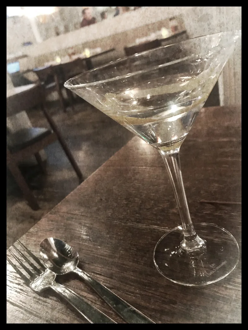 A partially filled martini glass is on a wooden table next to a spoon and fork. Background shows restaurant seating.