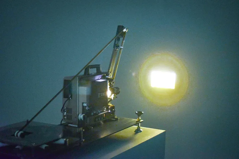 A film projector on a table projects light onto a wall, with a bright square of light visible.
