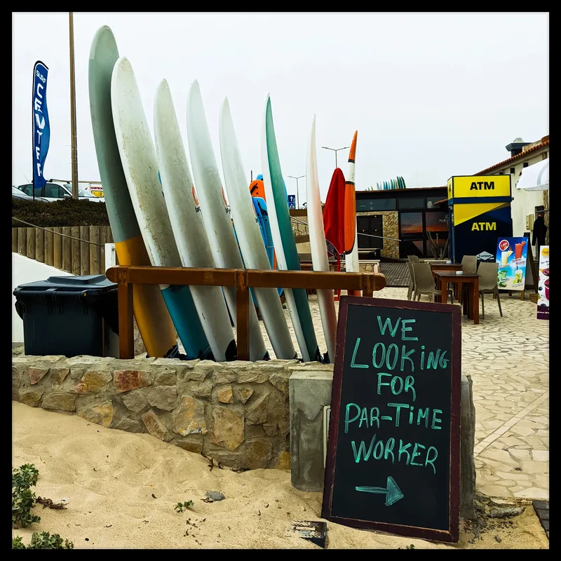 Row of surfboards propped against a wooden railing near a sign advertising part-time job, with various stalls behind.