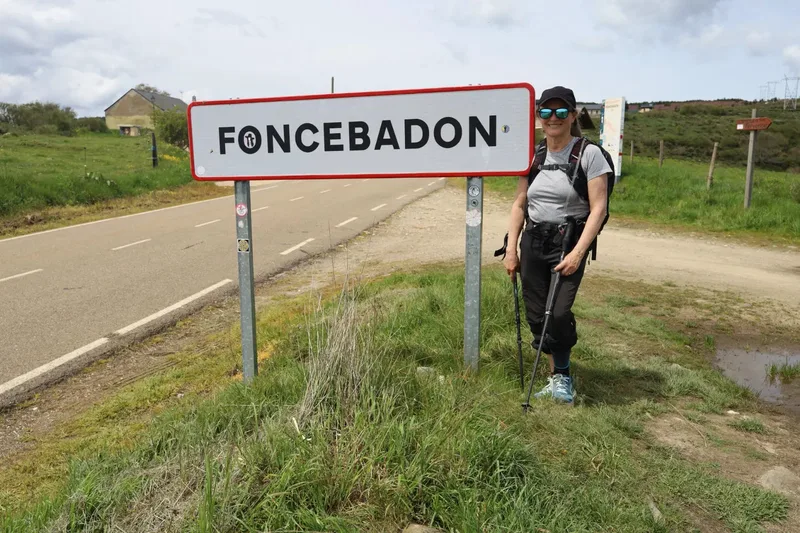 A person stands next to a Foncebadon sign beside a road, with grassy fields and a cloudy sky in the background.