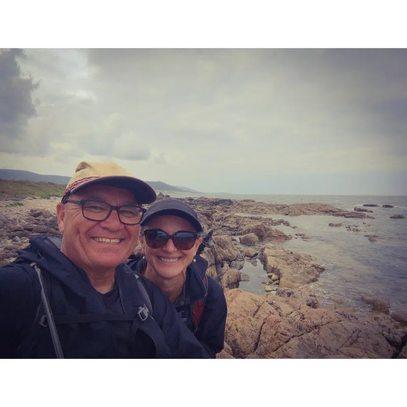 A man and a woman smile for a selfie on a rocky shoreline under a cloudy sky.