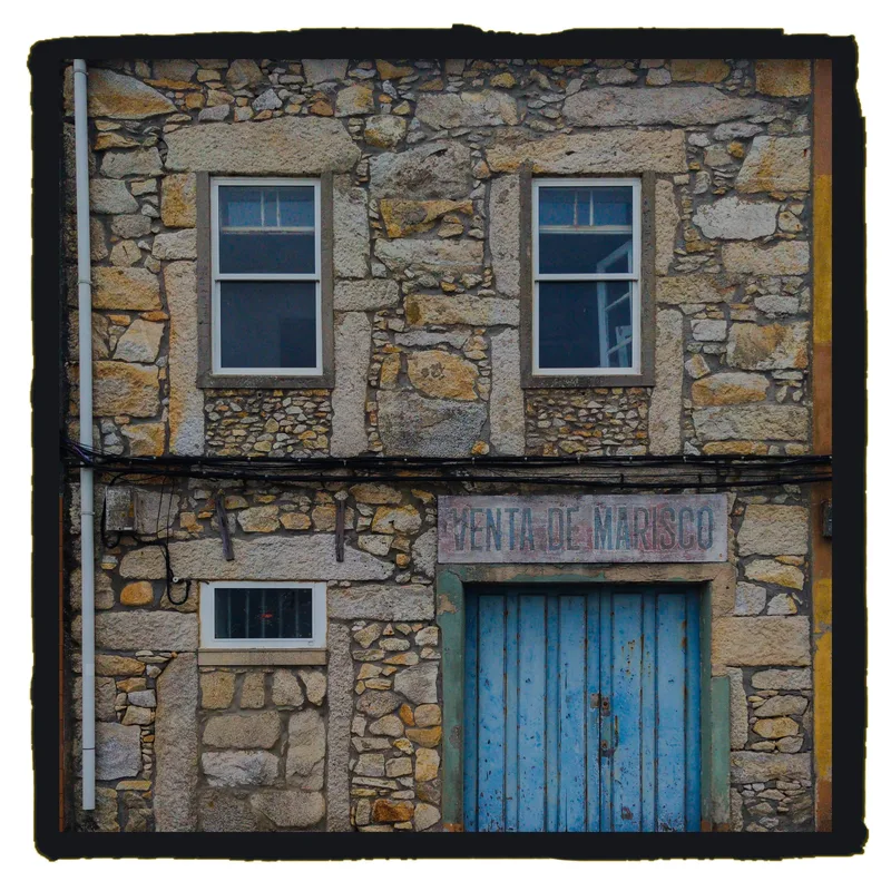 Stone building facade with two windows, a blue door, and a sign that reads "VENTA DE MARISCO.