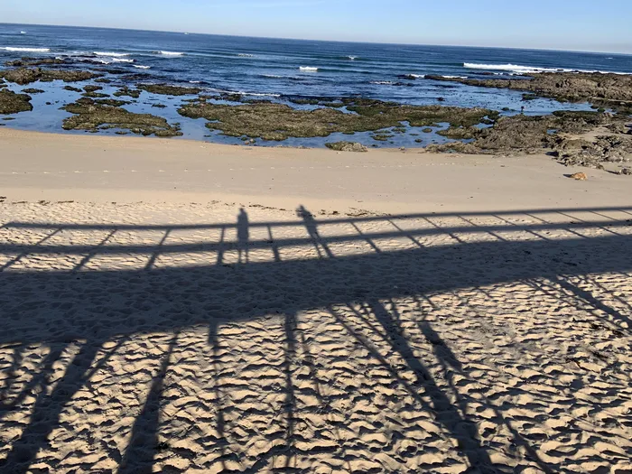 Beach scene with shadows cast on the sand, rocky coastline, and ocean waves in the background.