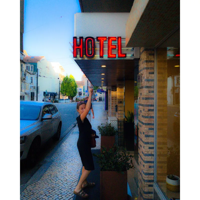 A woman in a black dress points to a "HOTEL" sign outside a building on a street with parked cars and trees.