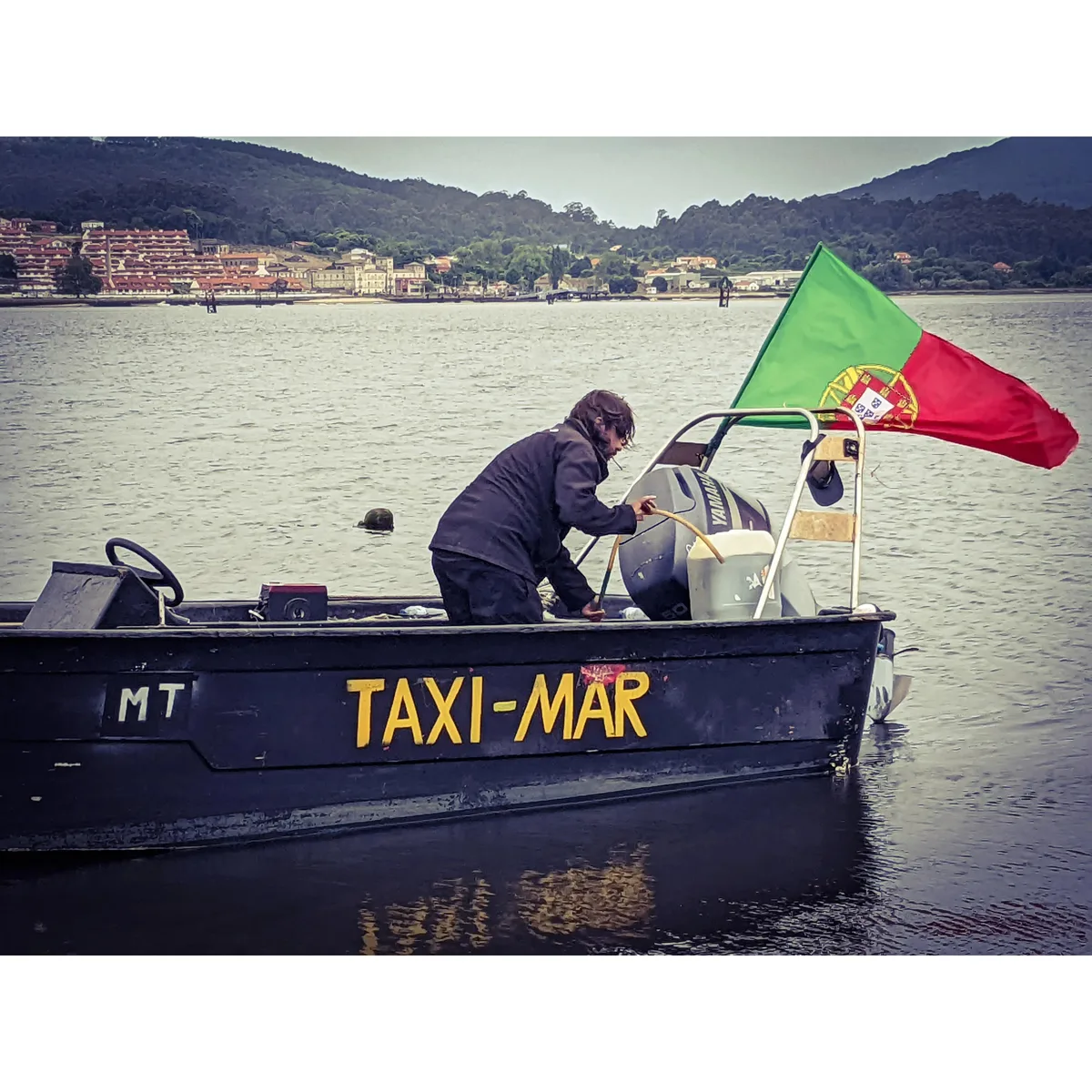 A man in a dark jacket refuels the motor of a Taxi-Mar boat flying a Portuguese flag, with a Spanish town on the shore behind