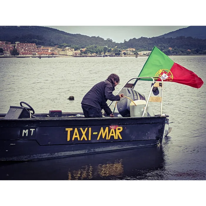 A man in a dark jacket refuels the motor of a Taxi-Mar boat flying a Portuguese flag, with a Spanish town on the shore behind