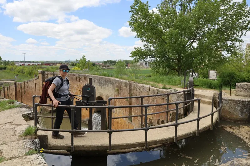 A person stands on a bridge near a water lock with greenery and a cloudy sky in the background.
