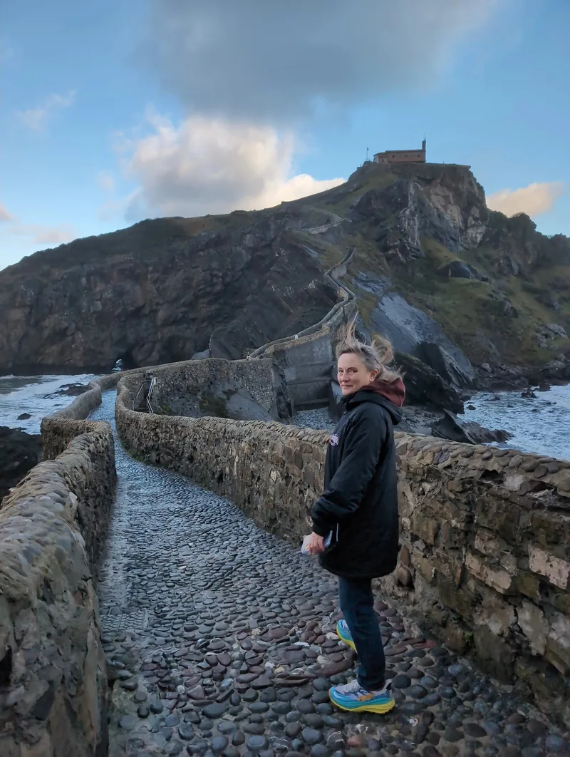 A woman stands on a stone pathway leading up a cliff with a building at the top, surrounded by rocky coast and ocean.