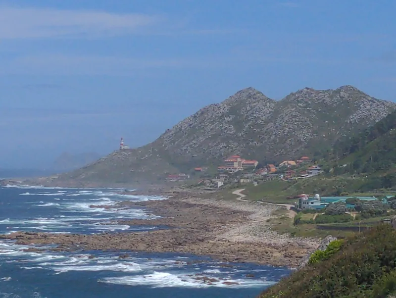Coastal landscape with a lighthouse, rocky shore, and green hills under a blue sky.