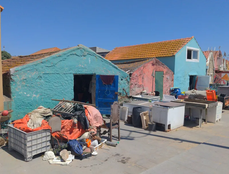 Colorful, weathered fish huts with clutter, including nets and containers, under a clear blue sky.