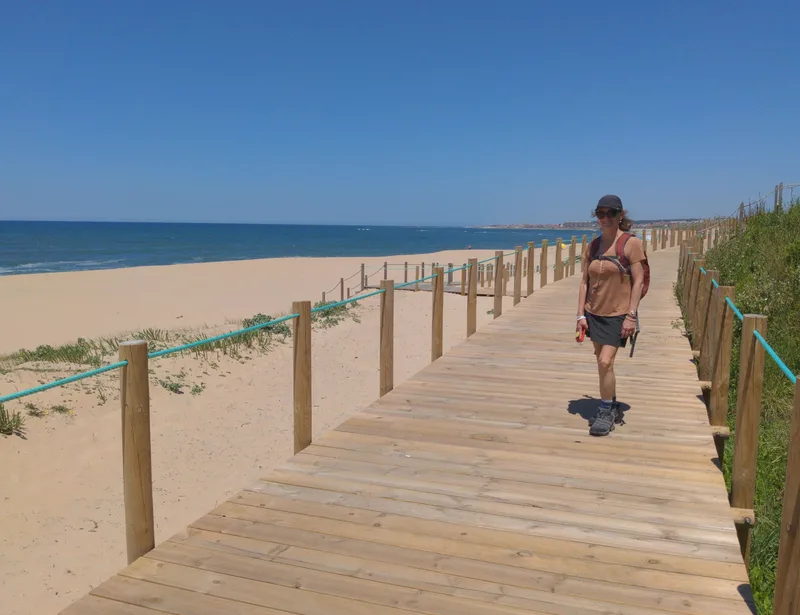 A person walks on a wooden boardwalk by the beach with sand and sea in the background under a clear blue sky.