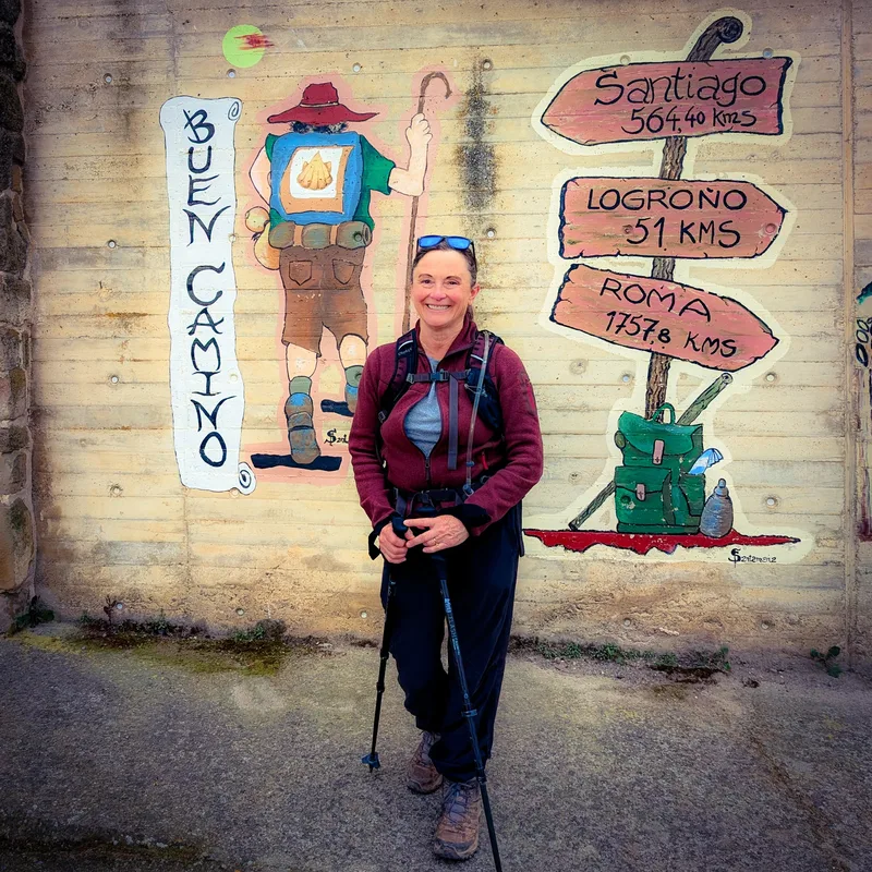 A woman with a backpack stands in front of a mural showing a hiker and distance signs to Santiago, Logroño, and Roma.