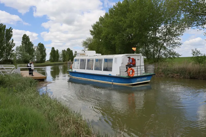 A blue and white boat navigates a calm canal near a wooden platform where two people stand. Trees and clouds are visible.