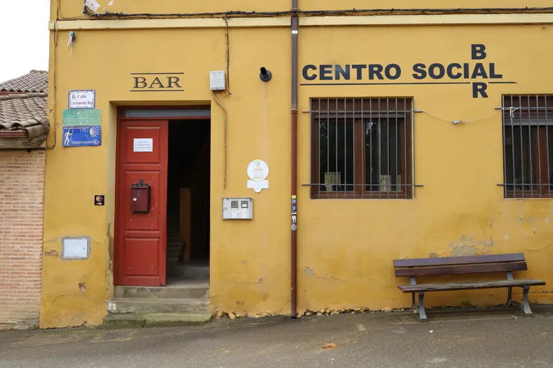 A yellow building with a red door, labeled "BAR" and "CENTRO SOCIAL," featuring windows and a bench outside.