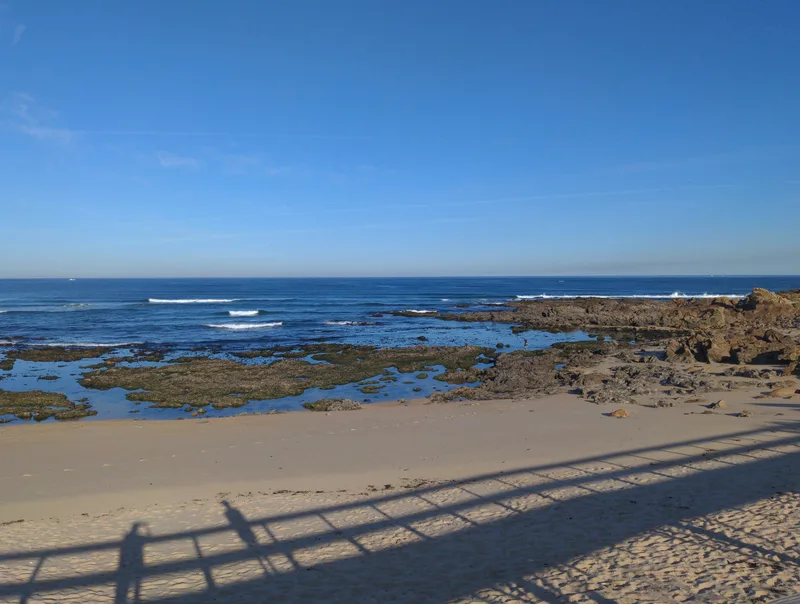 A sandy beach with rocky tide pools, clear blue sky, and gentle waves on the ocean. Shadows of a railing visible.