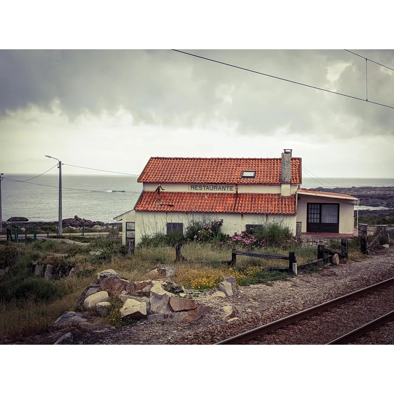 A small restaurant with a red-tiled roof near the ocean, surrounded by grass and rocky terrain.