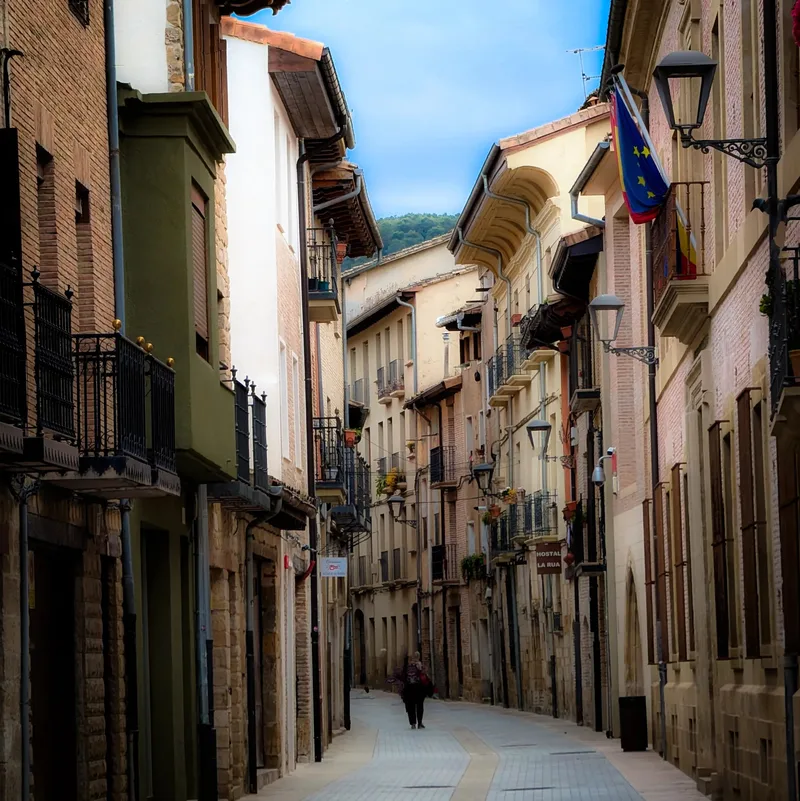 Narrow street lined with tall buildings, balconies, and a person walking in the distance under a blue sky.