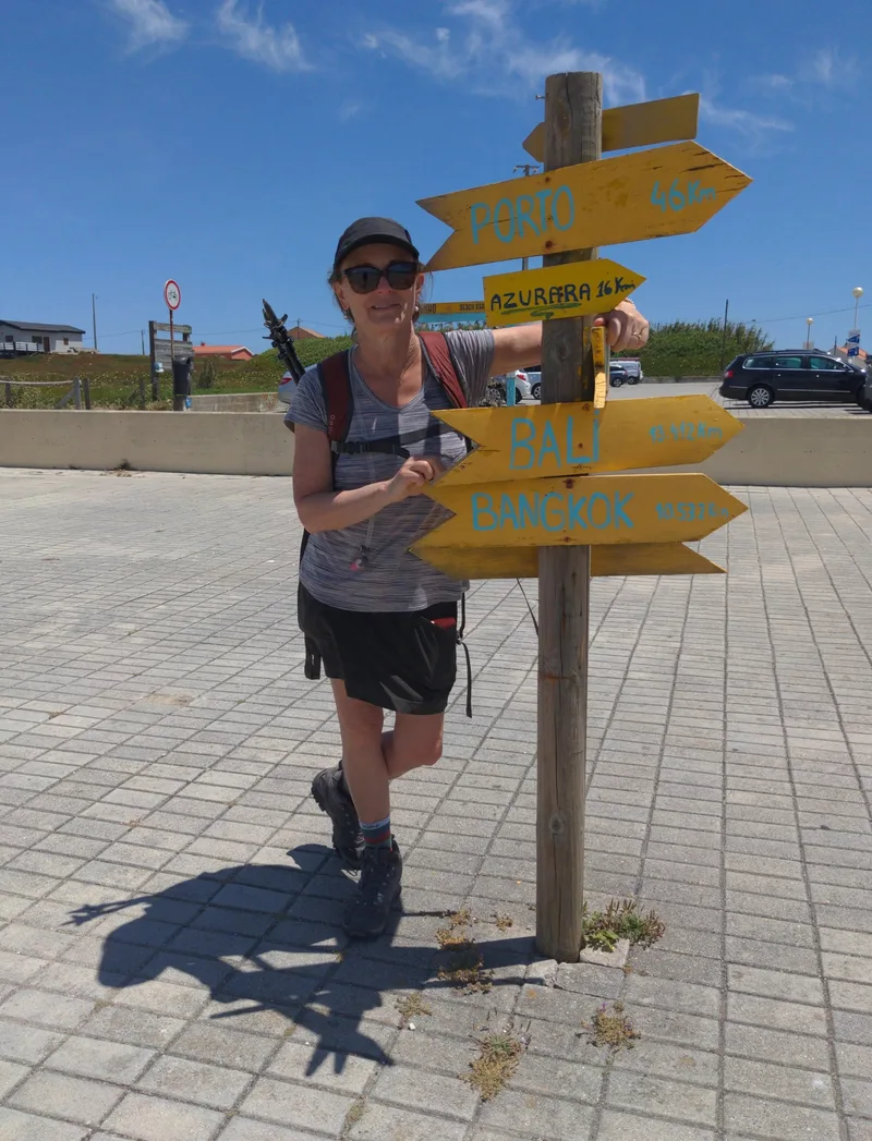 A woman in sunglasses stands beside a wooden signpost with directional arrows indicating distances to various places.