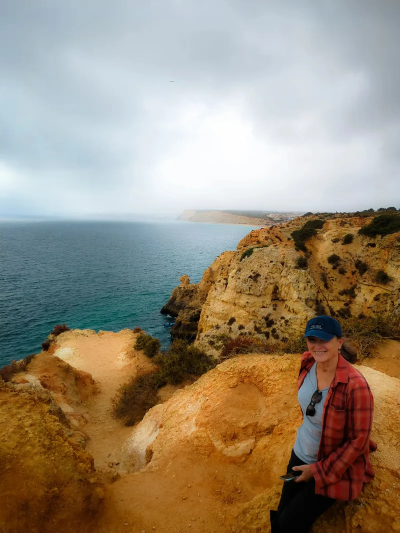 A person in a cap and flannel shirt sits on a rocky cliff overlooking a cloudy ocean landscape.