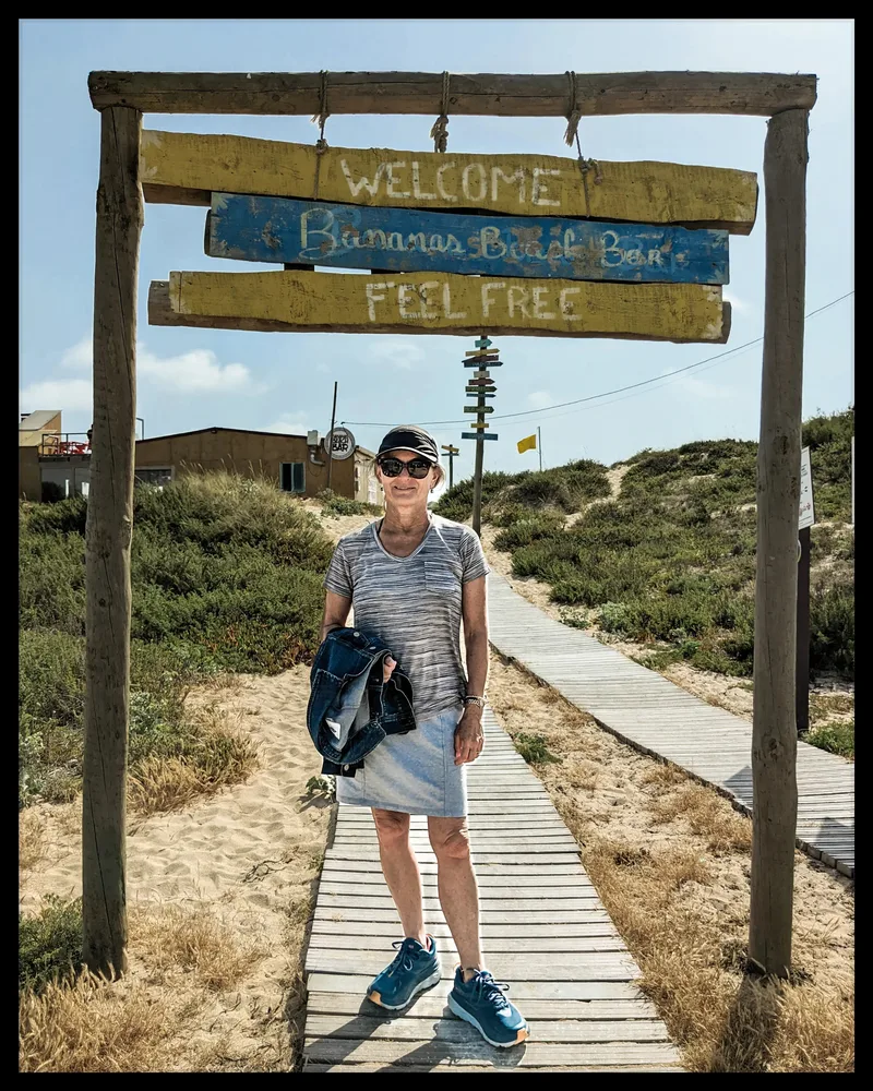A woman stands at a wooden sign reading "Welcome to Bananas Beach Bar," wearing a striped shirt and denim jacket.
