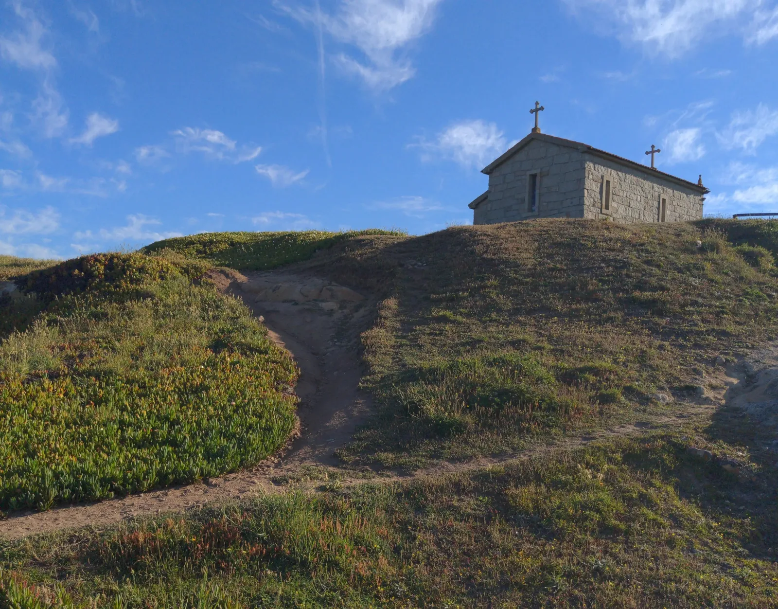 A stone church with three crosses on a hillside, surrounded by greenery and a blue sky with clouds.
