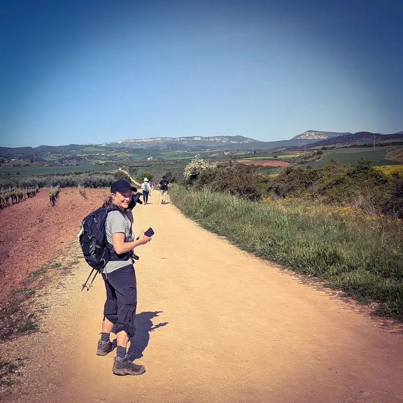 A person with a backpack stands on a dirt path, smiling and holding a phone, with hills and other hikers in the background.