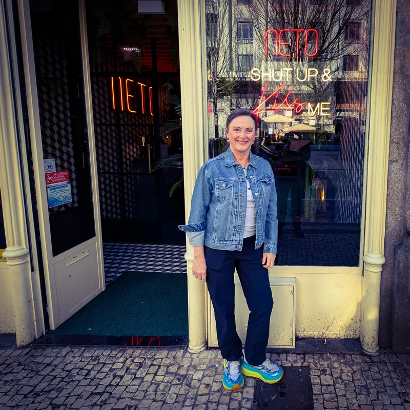 Woman in a denim jacket and pants stands at the entrance of a restaurant with neon signs.