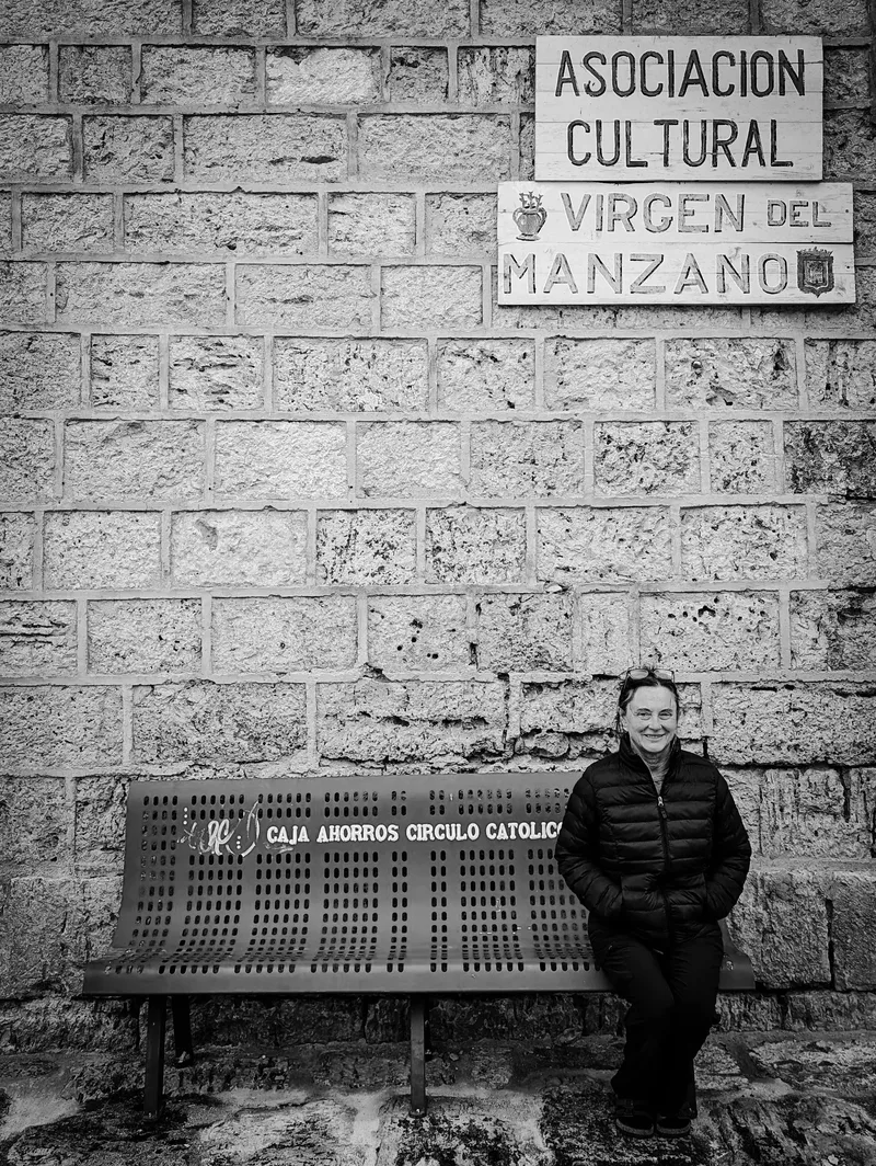 A woman in a black jacket sits on a bench against a stone wall with signs above it.