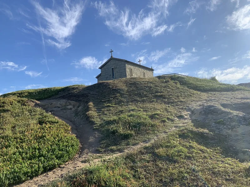 A stone church sits atop a grassy hill under a blue sky with clouds. A dirt path leads up to the church.