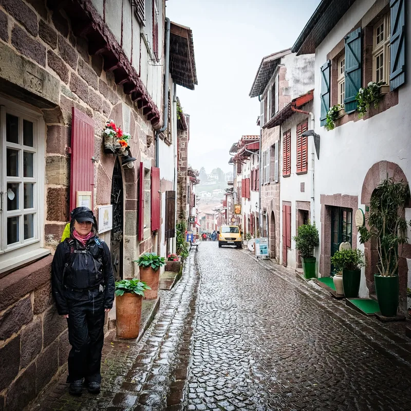 A narrow cobblestone street with historic buildings, a person in rain gear, and a yellow vehicle in the background.