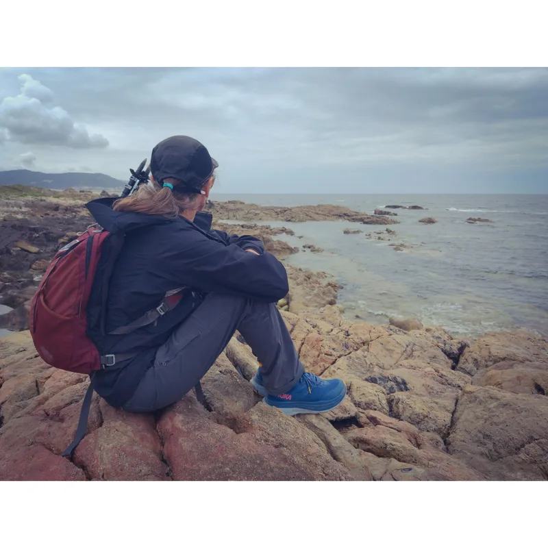 A person sits on rocky shore, facing the sea, wearing a backpack, dark clothing, and blue shoes under a cloudy sky.