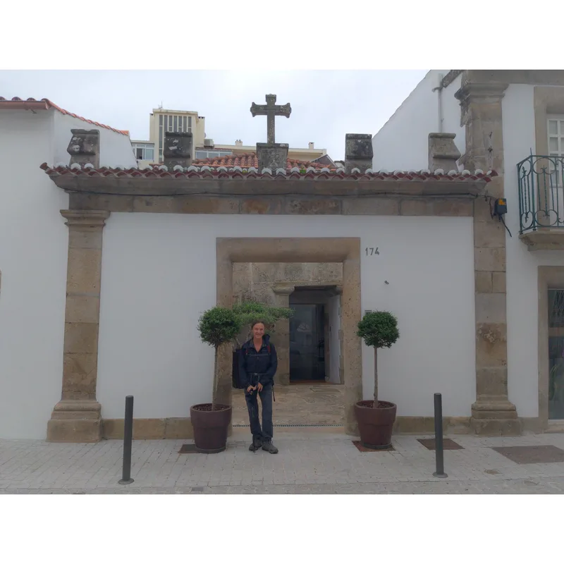 A person stands between two small trees in planters at the entrance of a white building with stone accents.