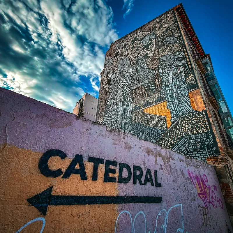A large mural depicting two figures on a building wall, with a pink wall displaying the word "CATEDRAL" and an arrow.