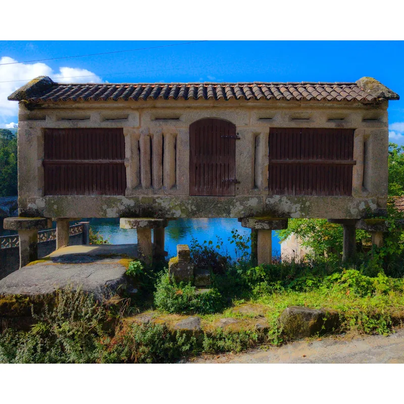 A stone granary on pillars beside a body of water, with wooden doors and a tiled roof, surrounded by greenery.