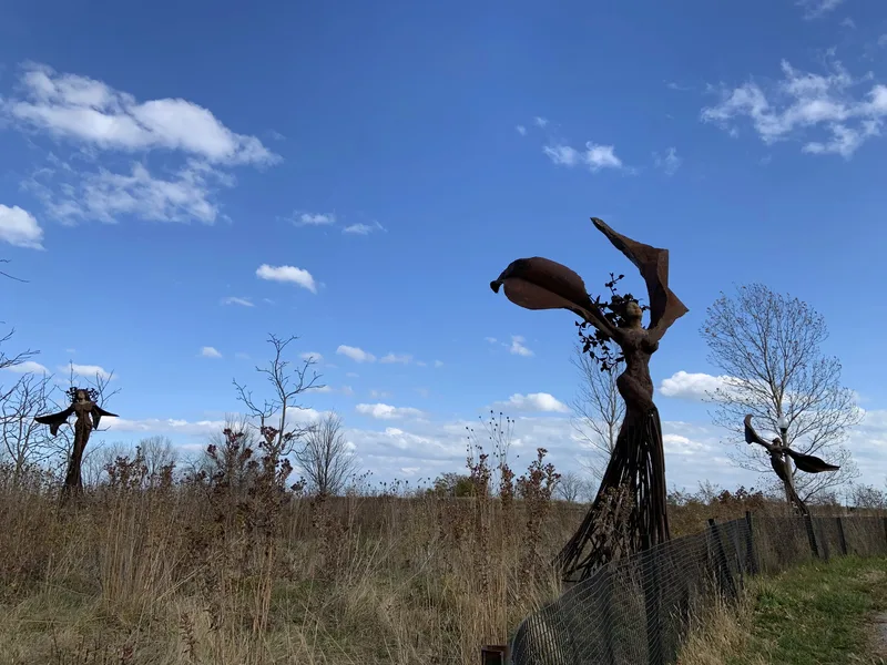 Three rusted metal sculptures resembling figures with wings in a grassy field under a blue sky with clouds.