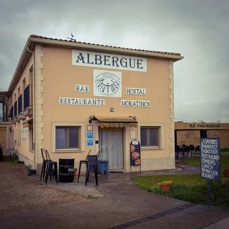A two-story building with "ALBERGUE" and "HOSTAL MORATINOS" signs, outdoor seating, and a menu board.