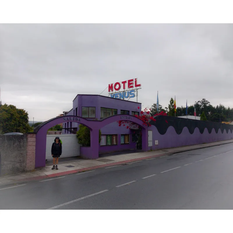 A purple motel building with a "MOTEL VENUS" sign; a person stands by an entrance on a cloudy street.