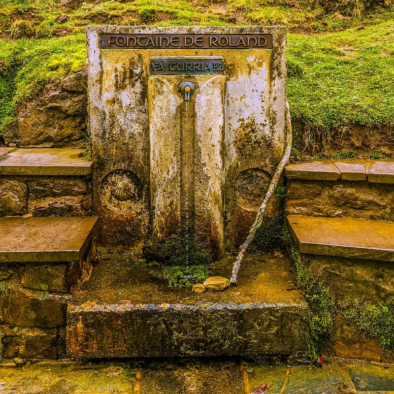 A stone fountain with a spout, labeled "Fontaine de Roland," set against a grassy hillside and stone steps.
