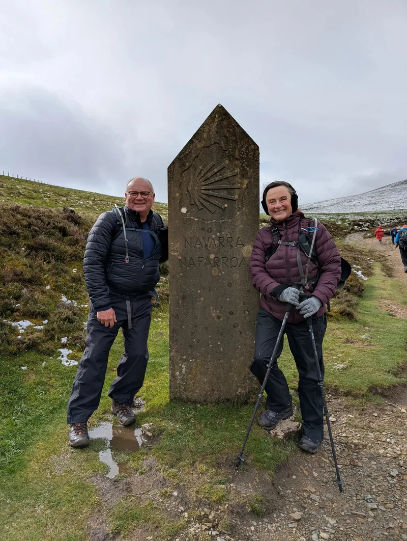 Two hikers stand beside a stone marker inscribed with "Navarra" in a grassy, outdoor setting.