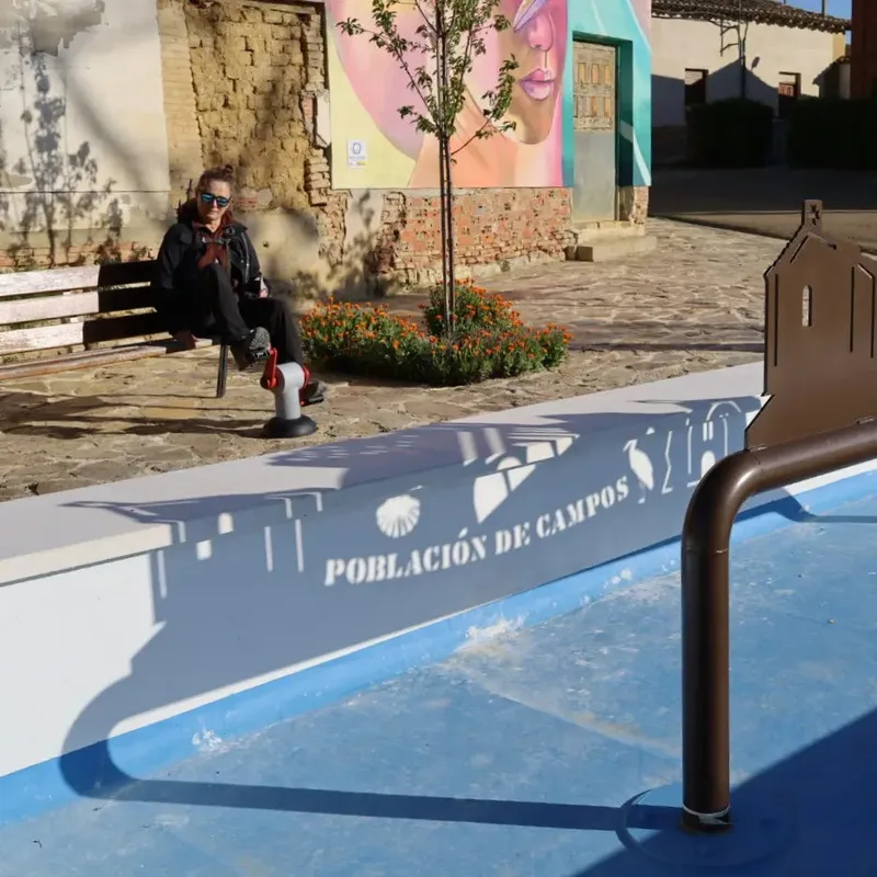 A person sits on a bench in a plaza with a mural, flowering plants, and a shadow depicting "Población de Campos.