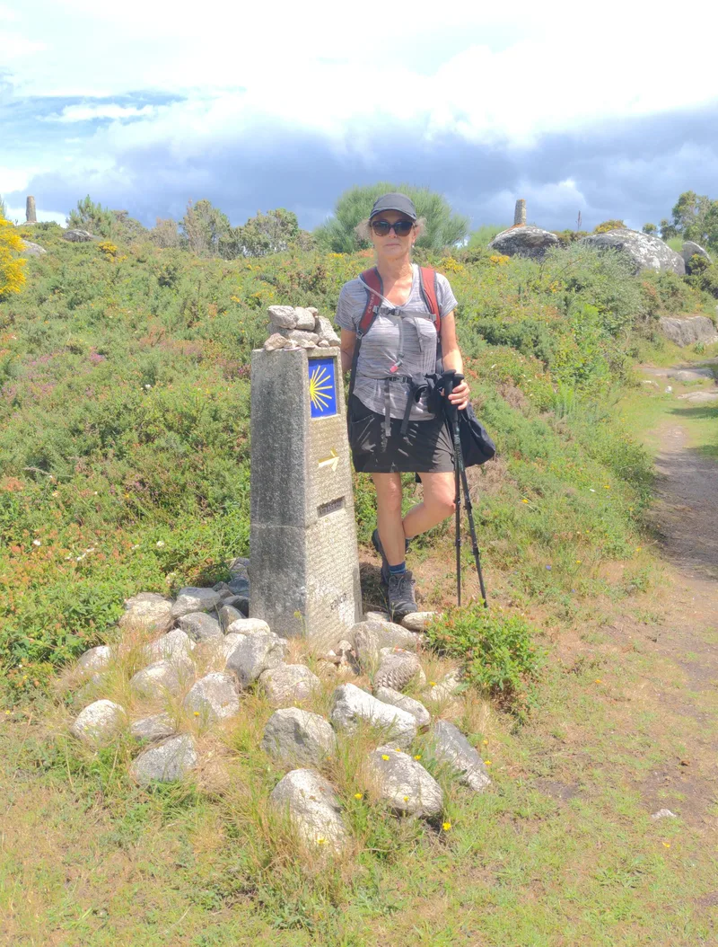 A woman stands beside a stone marker with a blue shell symbol, in a grassy area surrounded by rocks and vegetation.