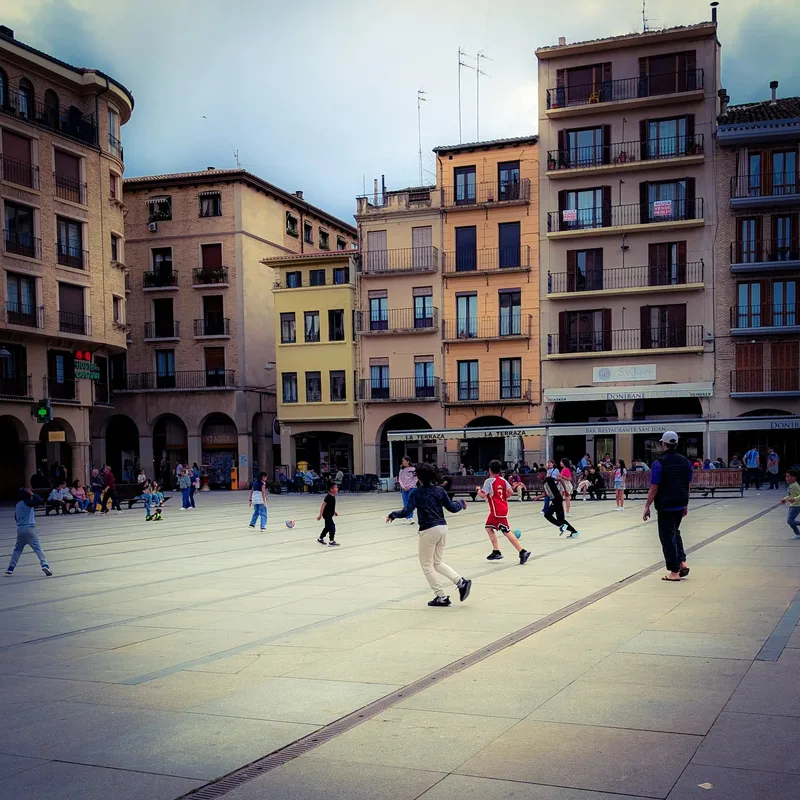 Children play soccer in a plaza surrounded by buildings and people sitting at outdoor cafes.