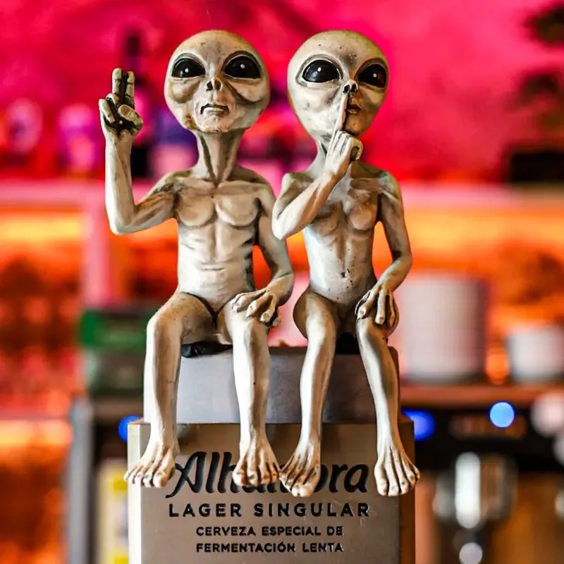 Two alien figurines sit on a pedestal, one making a peace sign and the other shushing, with a colorful backdrop.