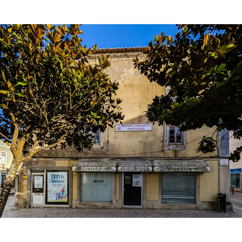 A building with a faded facade, featuring a sign for the "Partido Comunista Português," surrounded by trees.